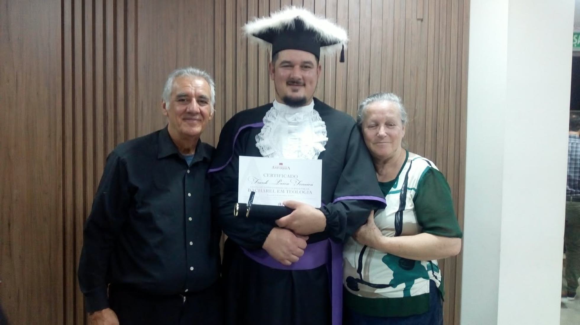 Sinval e uma mulher ao lado de um formando de beca, com o certificado nas mãos