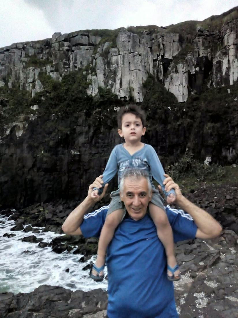 Um senhor com uma criança nos ombros diante de rochas e ondas