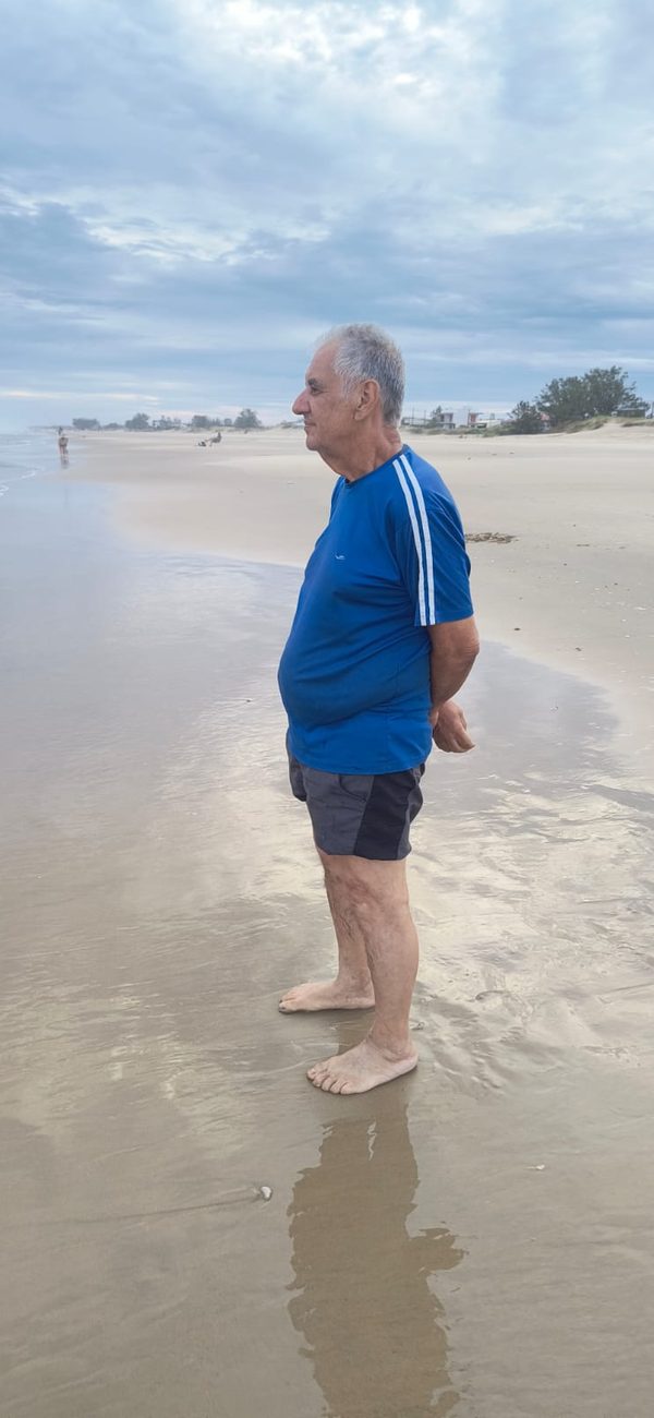 Sinval de perfil, de pé na beira da praia, olhando o mar