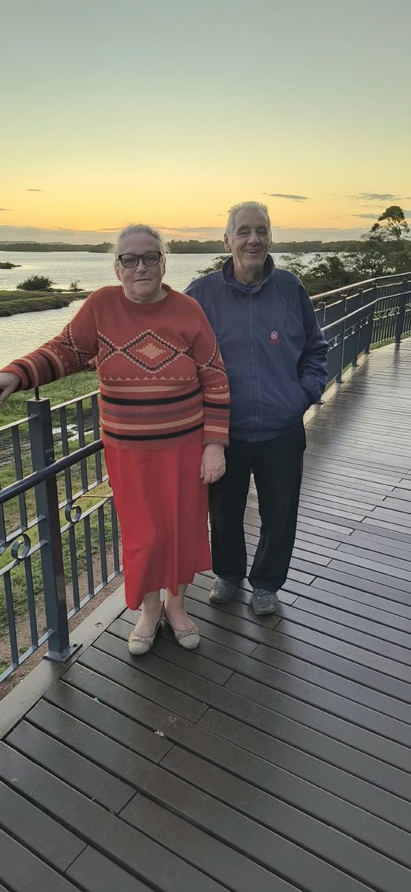 Sinval e uma mulher de pé num deque ao entardecer