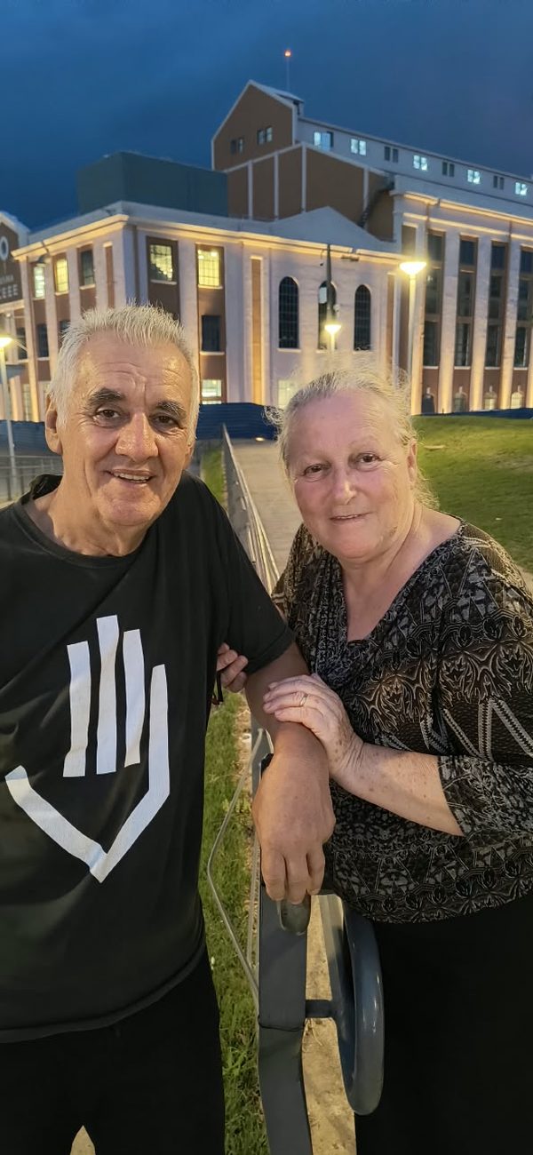 Sinval e uma mulher abraçados à noite, em frente a um prédio iluminado